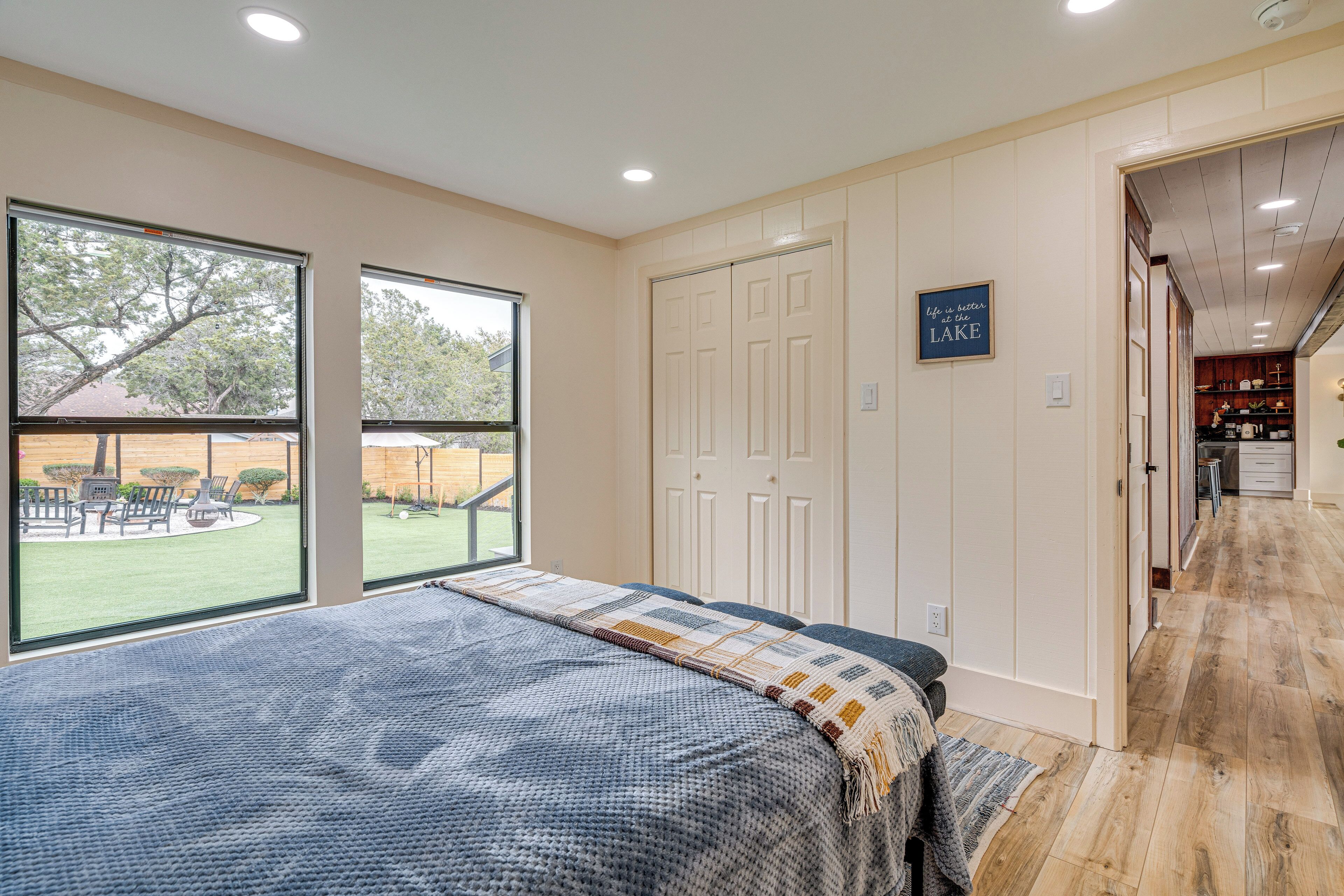 A bedroom with a large bed, a view of the backyard, and a hallway leading to a kitchen.