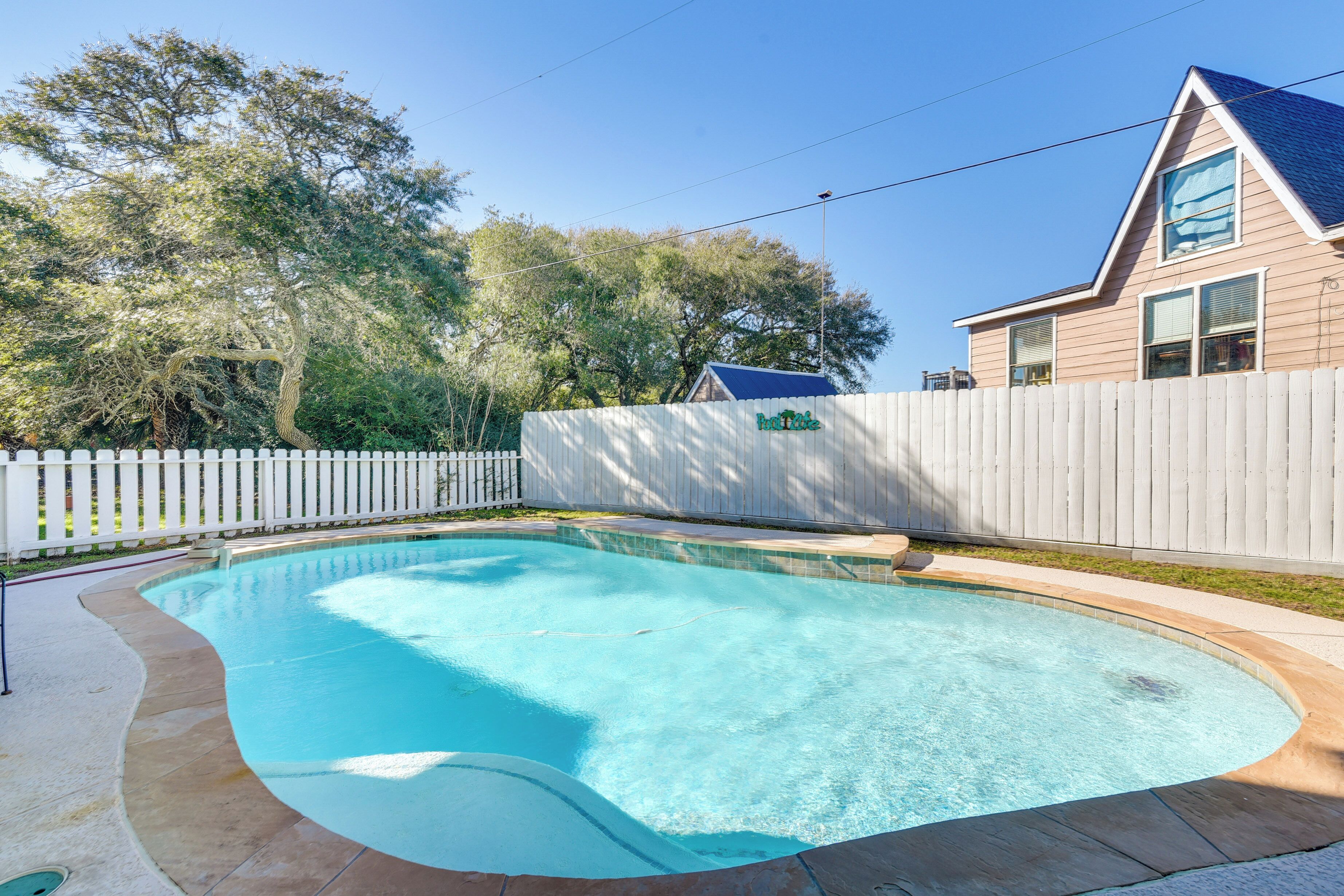 Una piscina de borde curvo, rodeada por una cerca de piquetes blancos y una casa con techo azul.