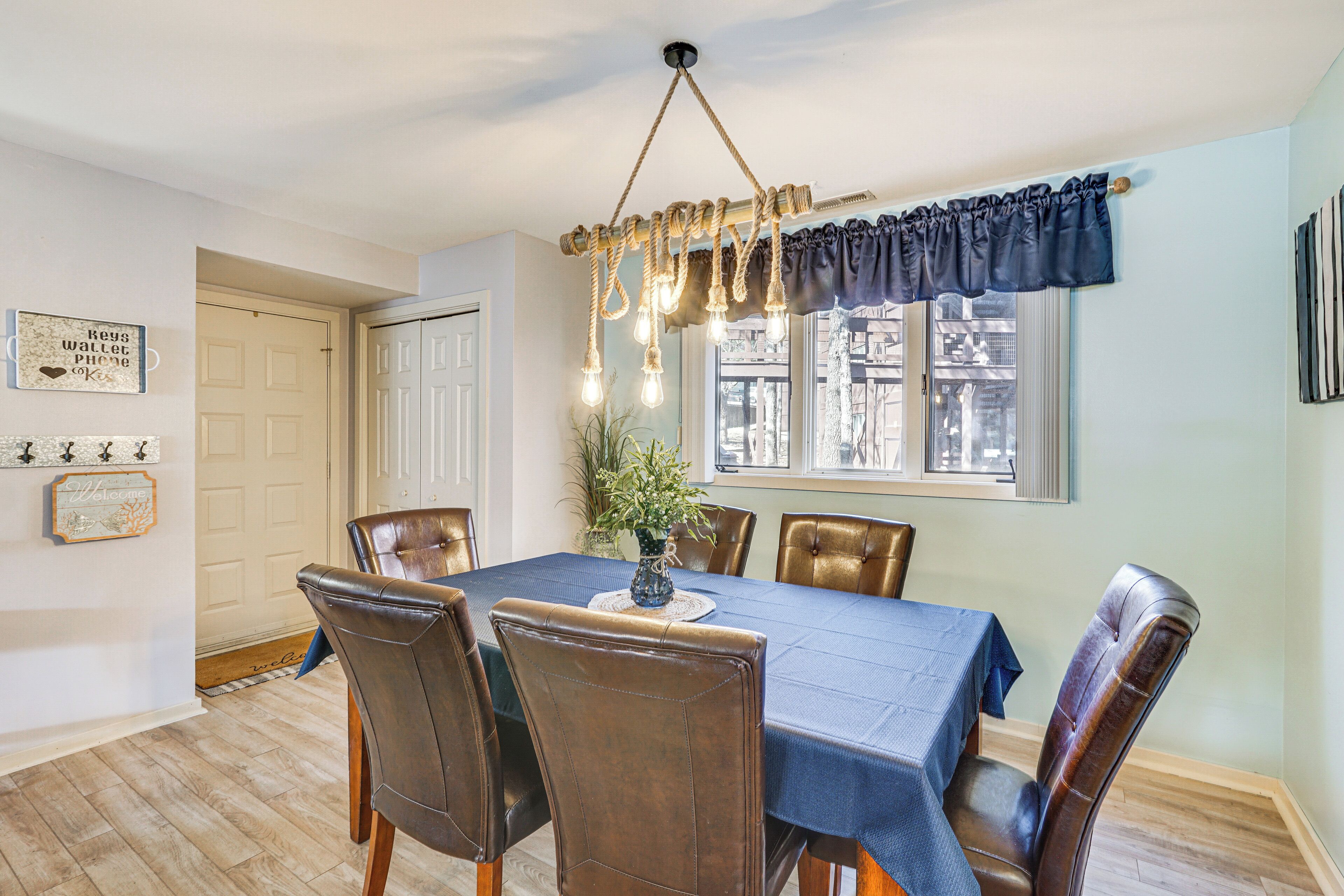 A dining room with a table set for six, surrounded by leather chairs, a chandelier, and a view of the city through a window with a blue valance.