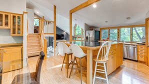 A spacious kitchen with wooden cabinets, a central island with bar stools, a dishwasher, and large windows offering views of the outdoors. - Calm in the Woods Fireplace Walk to Town (Suttons Bay)