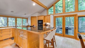 A modern kitchen with wooden cabinets, a large island, and a view of the forest through large windows. - Calm in the Woods Fireplace Walk to Town (Suttons Bay)