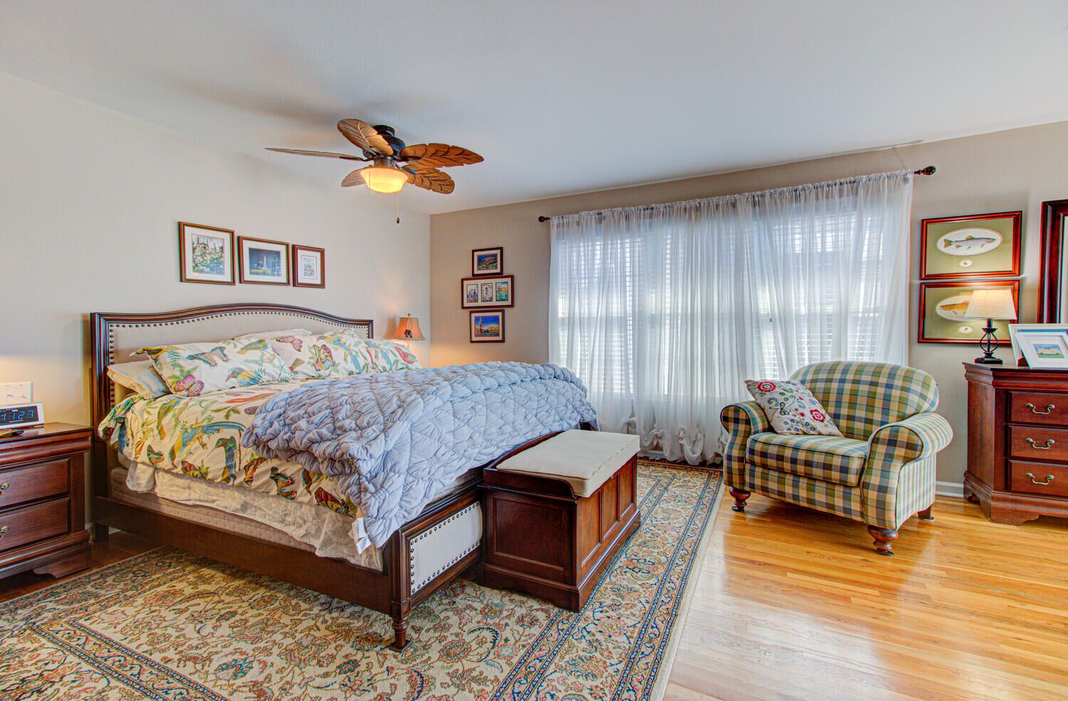 A bedroom with a bed, a chair, a nightstand, a bedside table, a rug, a ceiling fan, and a window with curtains.