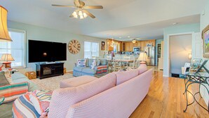 A spacious living room with a large TV, a ceiling fan, and a dining area with a kitchen in the background.