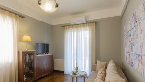 A living room with a sofa, a television, a cabinet, a table with a bottle and glasses, and a large window with curtains. - Le Boulevard (Corfu)