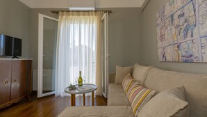 A living room with a sofa, a television, a small table with a bottle and glasses, and a window with curtains. - Le Boulevard (Corfu)