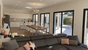 Ein modernes Wohnzimmer mit einer grauen Eckcouch, einem Essbereich mit Holztisch und -stühlen sowie einer Küche mit weißen Schränken.
