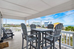 A covered outdoor deck with a table and chairs, overlooking a beach and houses.