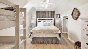 A bedroom with a bunk bed, a large bed with a headboard, a ceiling fan, and a nightstand with a lamp.