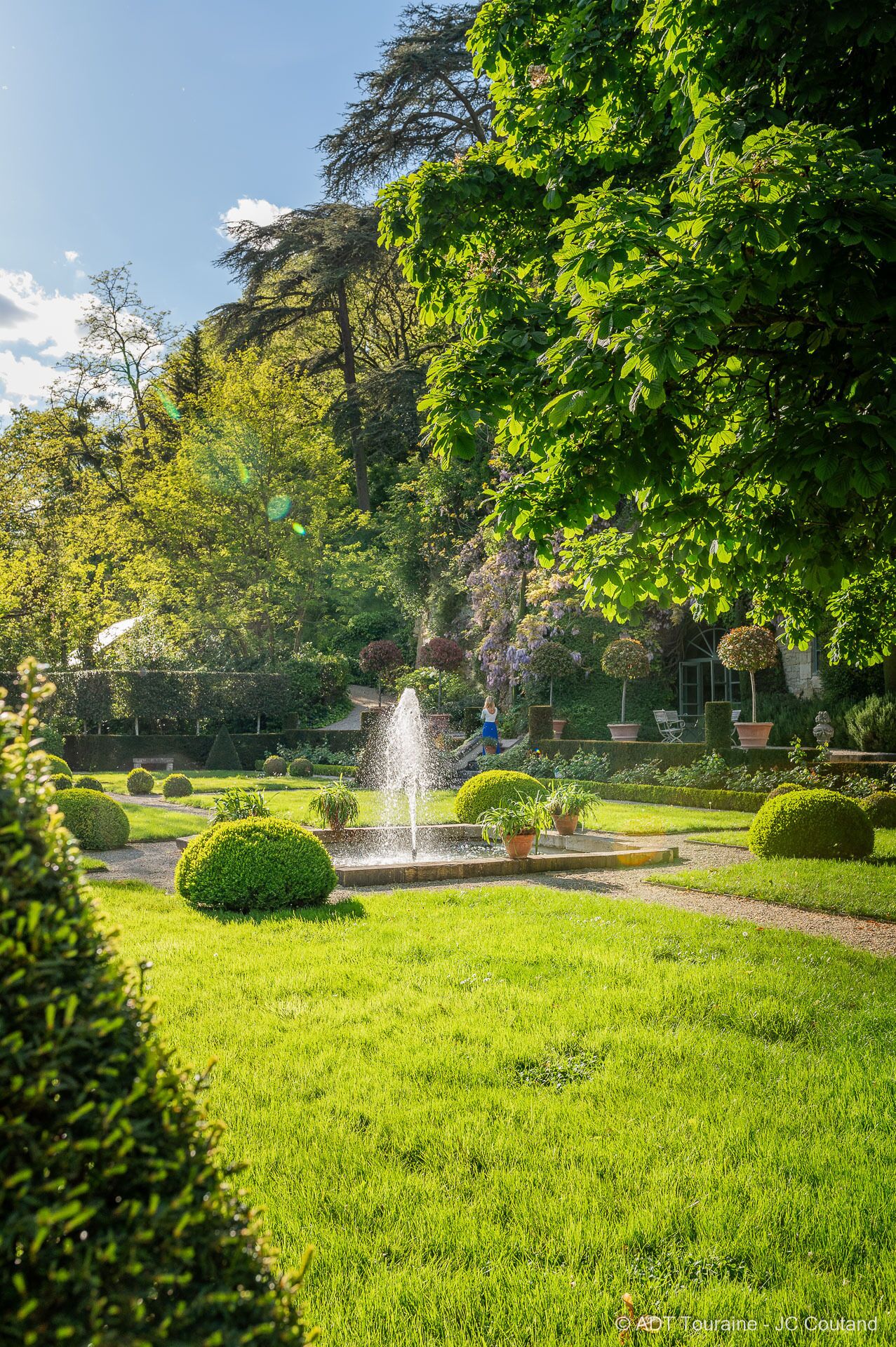 Un jardín bien cuidado con una fuente central, setos cuidadosamente recortados y una variedad de árboles.