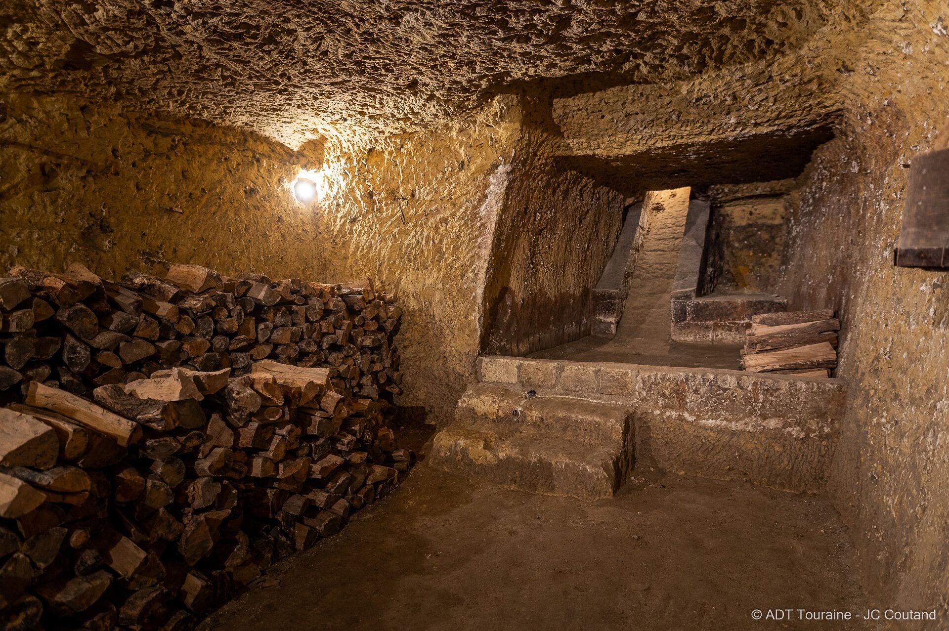 A dimly lit cave with rough stone walls, a pile of firewood, and a small window.