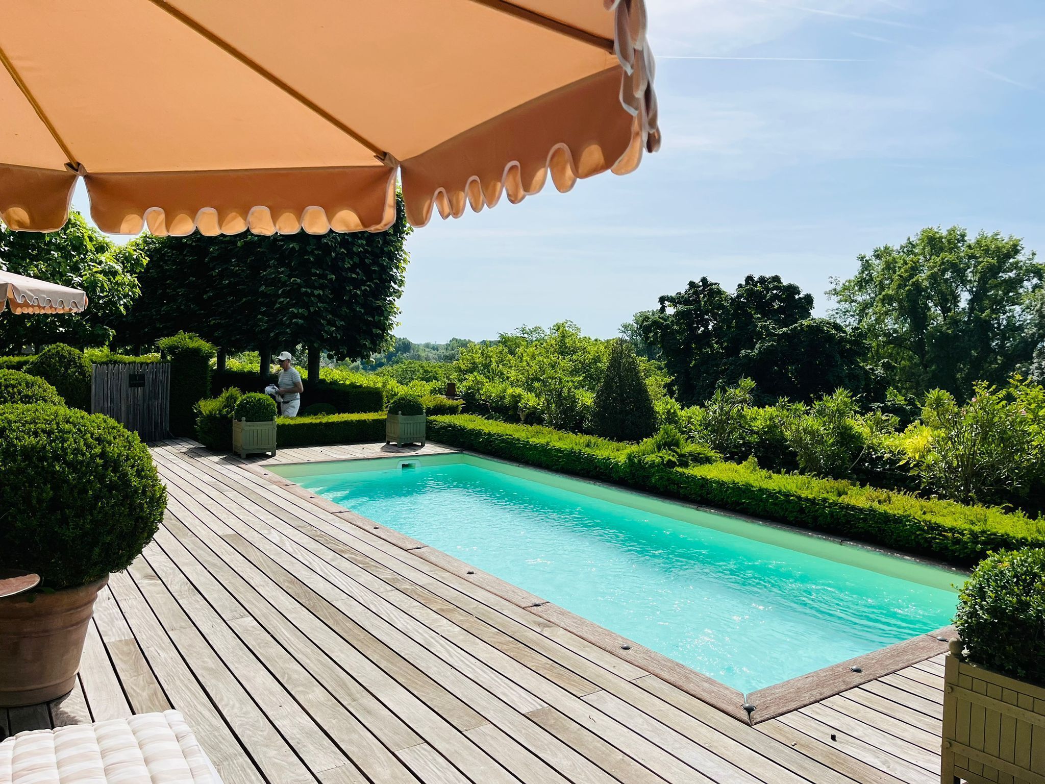 Une terrasse en bois avec une piscine, entourée de haies et d’arbres soigneusement entretenus.