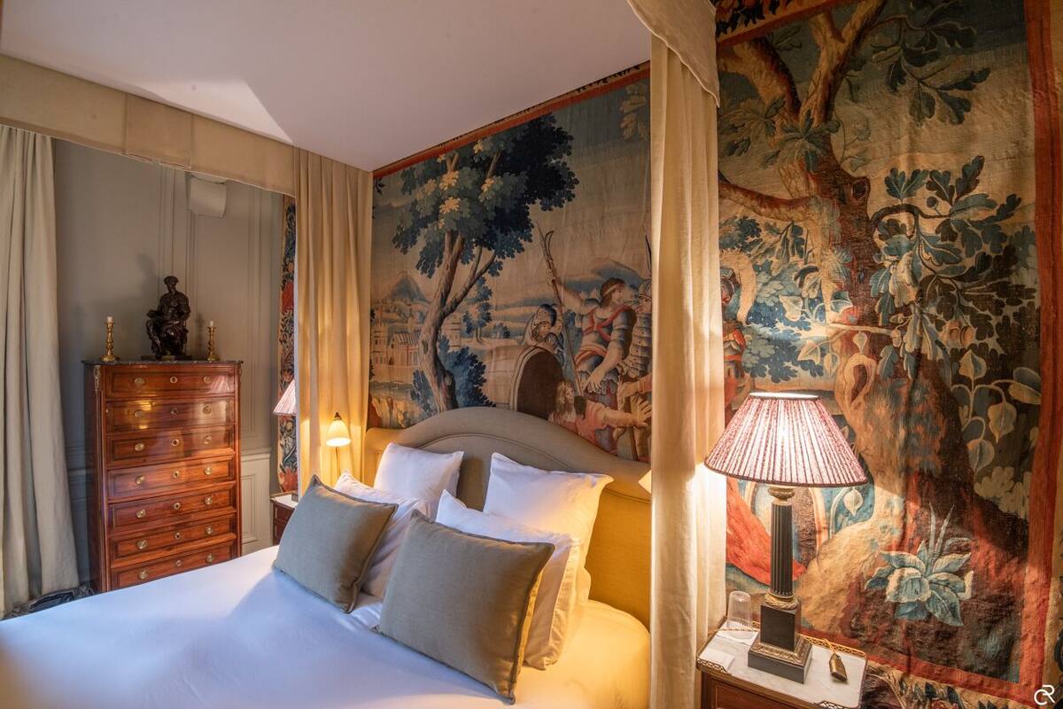 A hotel room with a large bed, a dresser, a lamp, and a tapestry on the wall.