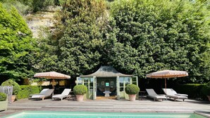 A pool with lounge chairs and umbrellas, surrounded by greenery and a small building with glass doors. - MANOIR DES BASSES RIVIERES (Rochecorbon)