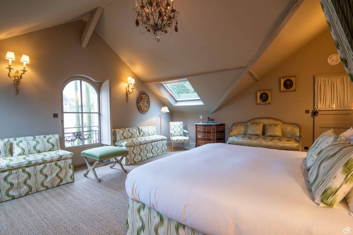 A bedroom with a large bed, a sofa, a chair, a small table, and a chandelier. There are two windows and a skylight.