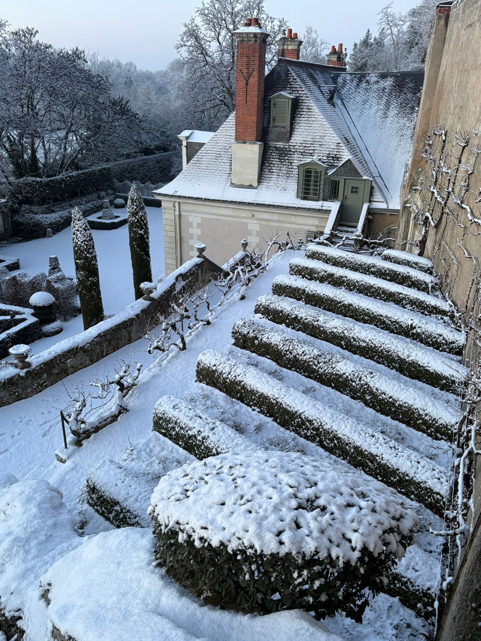 En snötäckt trappa leder upp till en byggnad med en tegelmurad skorsten, omgiven av snötyngda träd och buskar.