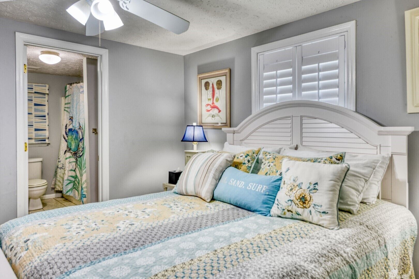 A bedroom with a bed, a nightstand, a lamp, a framed picture, and a window with shutters.