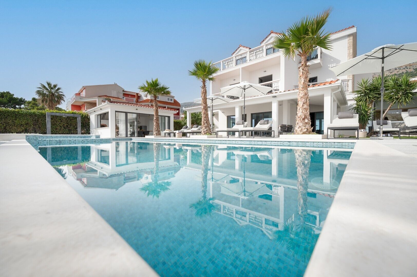 Een modern hotel met een groot zwembad, palmbomen en een helderblauwe lucht.