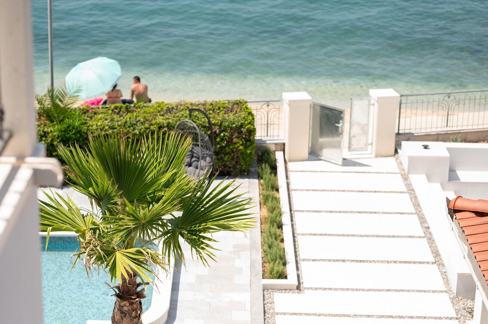 A pool area with a palm tree, a person under a blue umbrella, and a view of the sea.