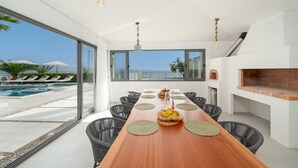 A modern dining area with a long wooden table, black wicker chairs, and a view of the sea through large windows.