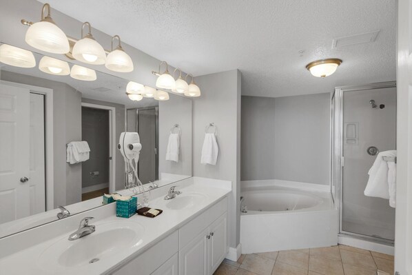 A bathroom with a large mirror, multiple light fixtures, a white sink, a vanity, a towel rack, a hair dryer, and a bathtub.