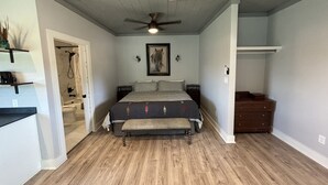 A bedroom with a wooden floor, a bed with a headboard, a ceiling fan, a painting on the wall, and a dresser. - Ranch Hand Cabin in Llano Texas  (Llano)