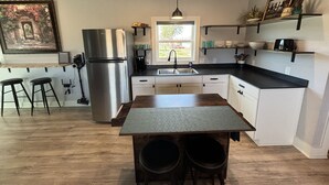 A modern kitchen with a stainless steel refrigerator, a wooden island with stools, and a window providing natural light. - Ranch Hand Cabin in Llano Texas  (Llano)