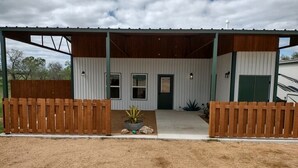 A single-story building with a covered porch, a green door, and a fenced driveway. - Ranch Hand Cabin in Llano Texas  (Llano)