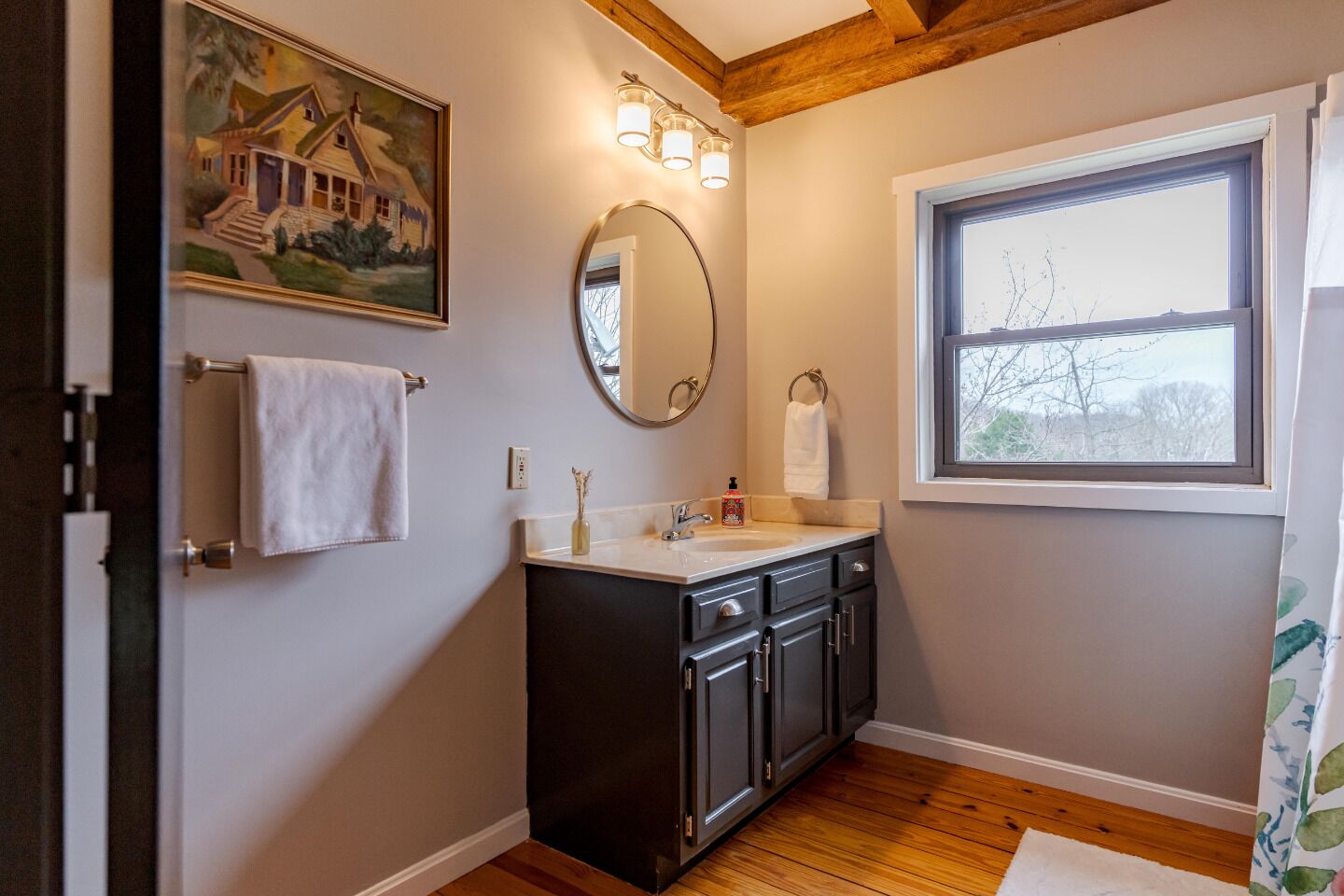 A bathroom with a wooden floor, a sink with a mirror, a window, and a painting on the wall.