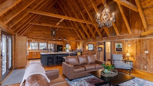 A spacious log cabin interior with a high ceiling, a central living area with a sofa and coffee table, a kitchen with modern appliances, and a dining area with a chair.