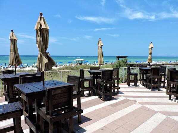 Un área de estar al aire libre con mesas y sillas de madera, sombrillas y vistas a la playa y al océano.