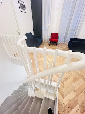 Uma escadaria com grades brancas leva a um quarto com padrão de piso em espinha de peixe, dois sofás de cor escura e uma cadeira vermelha.