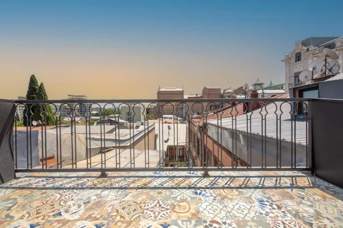 A rooftop terrace with a patterned floor, metal railing, and a view of surrounding buildings.