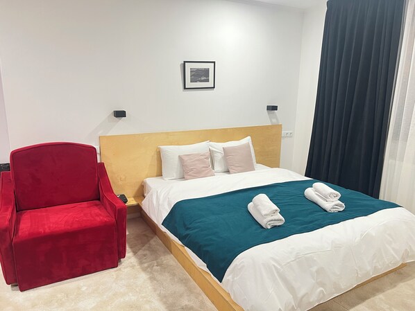 Habitación de hotel con cabecera de madera, un sillón rojo, cama con ropa de cama blanca y verde azulado, y un cuadro en la pared.