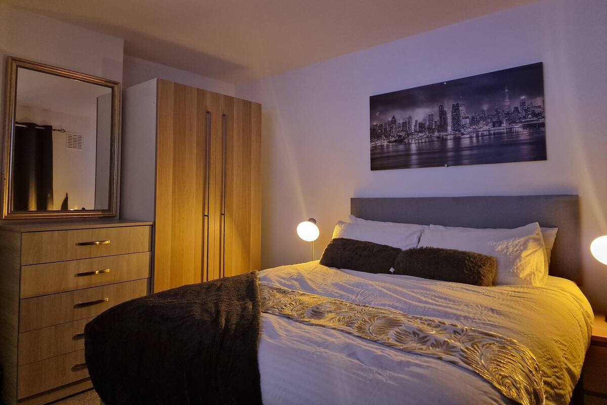 A bedroom with a bed, a wardrobe, a mirror, and a cityscape painting on the wall.