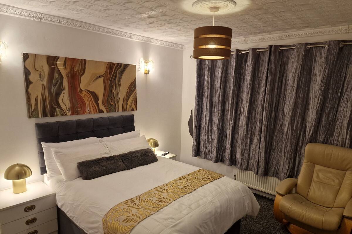A neatly made bed with white and gold bedding, a dark headboard, and a decorative wall piece above it.