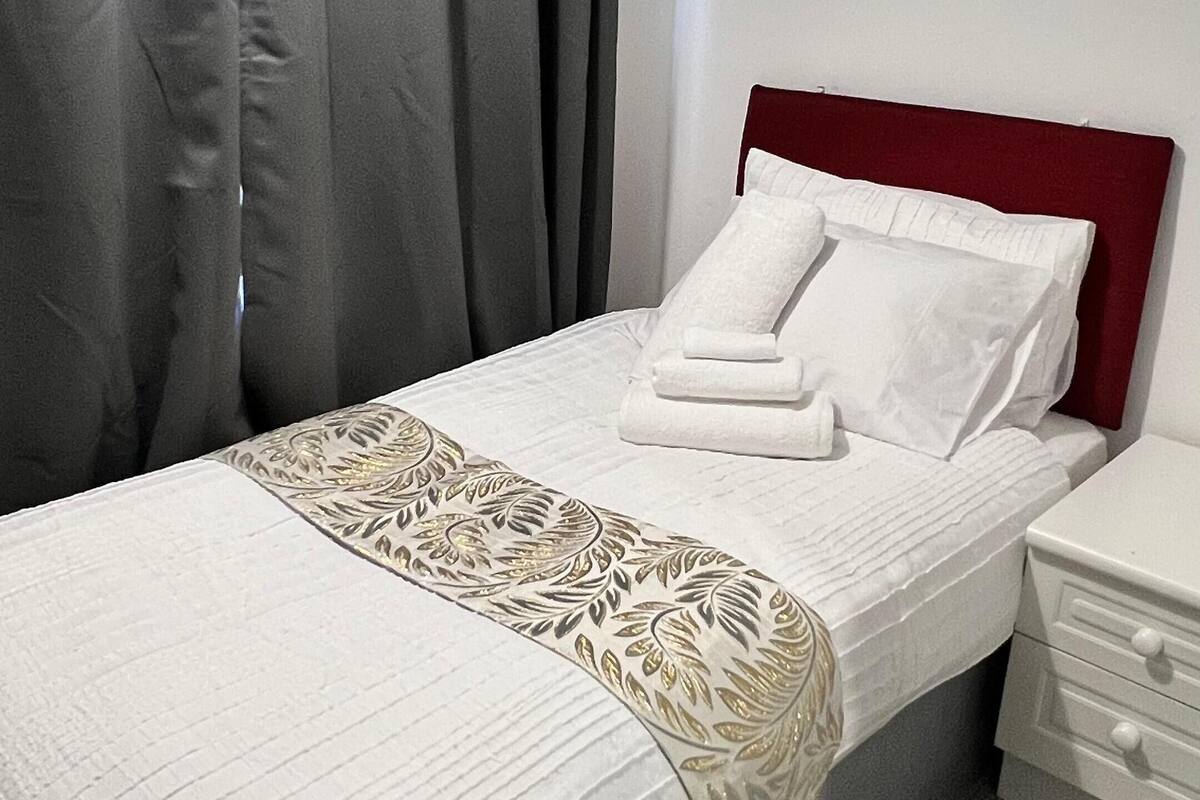 A single bed with a white bedspread and a decorative gold leaf pattern, a red headboard, white pillows, and a white nightstand with drawers.