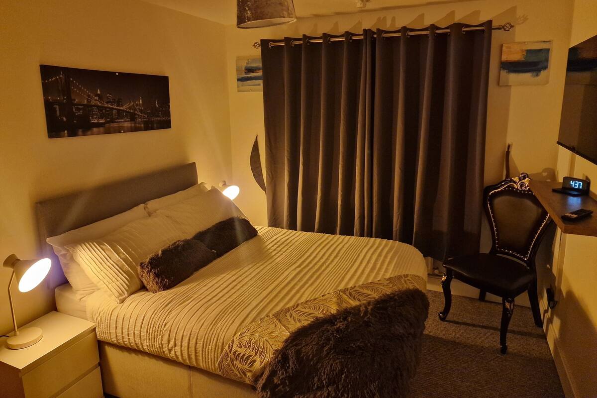 A hotel room with a bed, bedside lamps, a chair, a desk with a clock, a painting, and curtains.