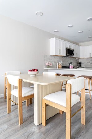 A modern kitchen with white cabinets, a central island, and a dining area with a light-colored table and wooden chairs.