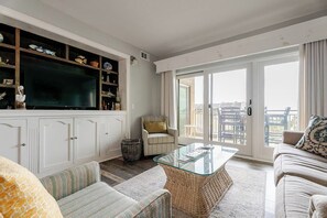 A spacious living room with a large flat-screen TV, wicker furniture, and a balcony with outdoor seating.
