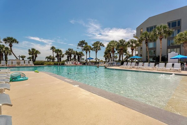 A swimming pool with lounge chairs and umbrellas, surrounded by palm trees and a building.