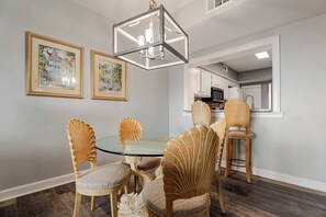 A dining area with a glass table surrounded by chairs with shell-like backs, a chandelier, and framed pictures on the wall.