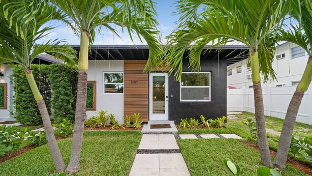 Une maison moderne avec un chemin pavé menant à l’entrée, entourée de palmiers et d’une pelouse bien entretenue.