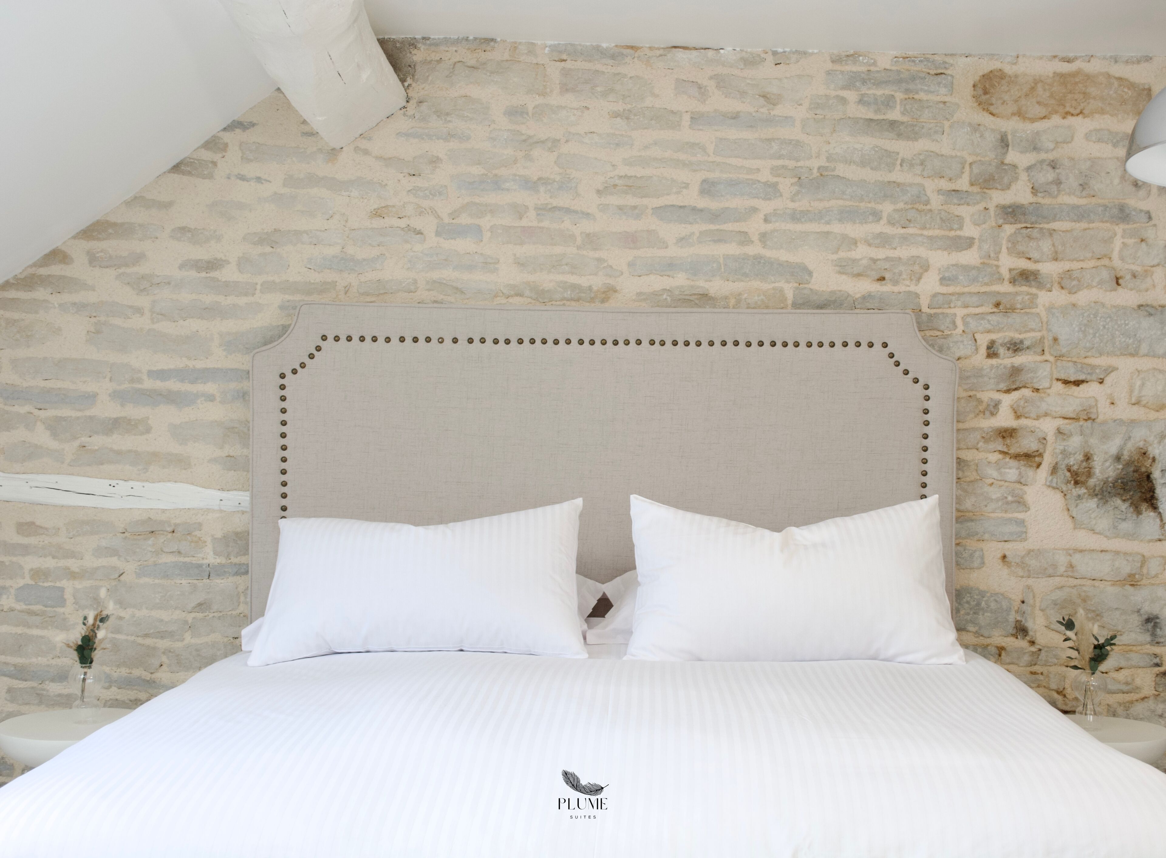 Un lit avec une tête de lit adossée à un mur en pierre, deux oreillers et un logo sur la couverture du lit.