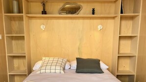 Una cama bien hecha con edredón blanco y dos almohadas, una mesita de noche con un florero, y estanterías empotradas de madera a ambos lados de la cama.