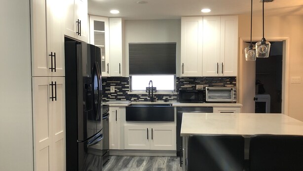A modern kitchen with white cabinets, a black refrigerator, a built-in oven, and a stainless steel sink. There is a glass dining table with two bar stools.