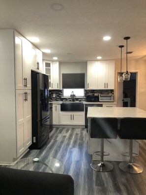 A modern kitchen with white cabinets, a black refrigerator, a built-in oven, and a stainless steel sink. There is a glass dining table with two bar stools. - Charming 3-bedroom house centrally located in Brandon, FL  (Brandon)