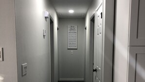 A narrow hallway with a wooden floor, white walls, and a ceiling vent. A sign with text is visible on the wall.