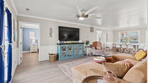 Een ruime woonkamer met een plafondventilator, een flatscreen-tv, een eethoek met een tafel en stoelen, en een gezellige zithoek met een bank en kussens.