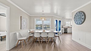 Een eetruimte met een grote houten tafel, zes stoelen, een kroonluchter en een grote klok aan de muur.