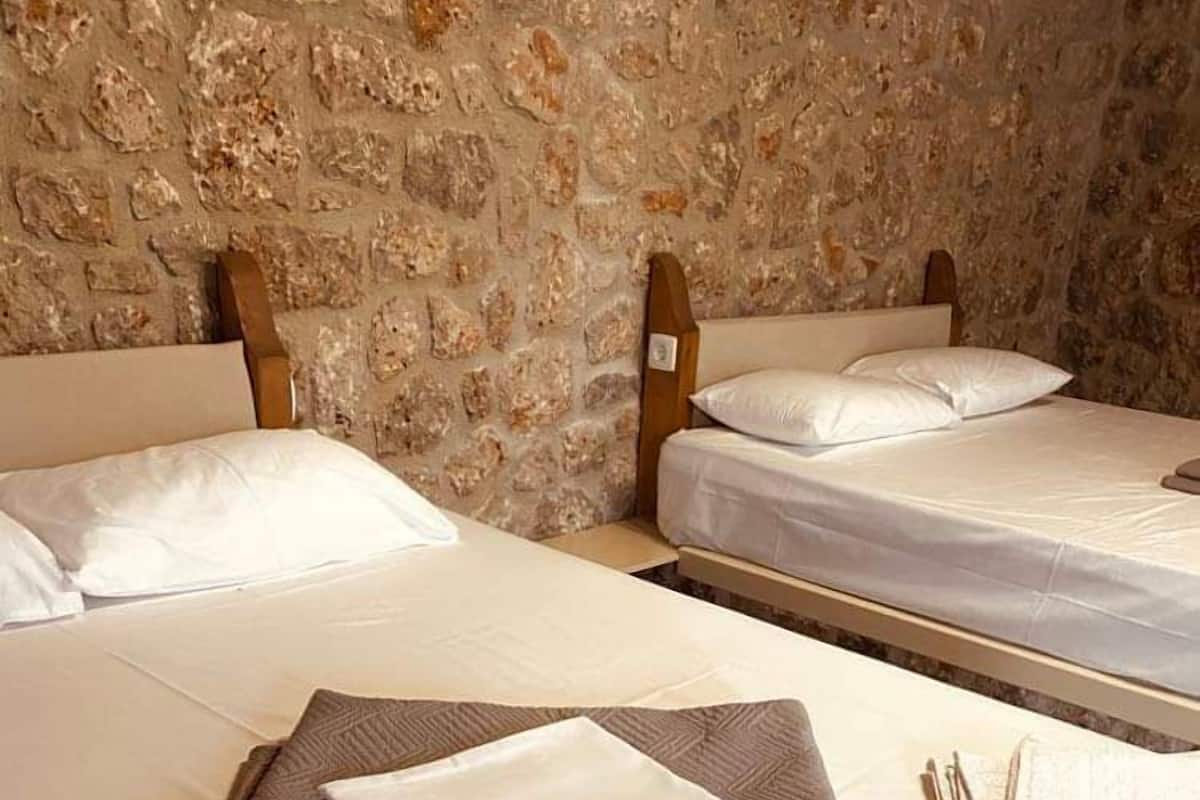 A hotel room with two beds, each with white linens and a folded towel on the bed. The room has a stone wall and wooden headboards.
